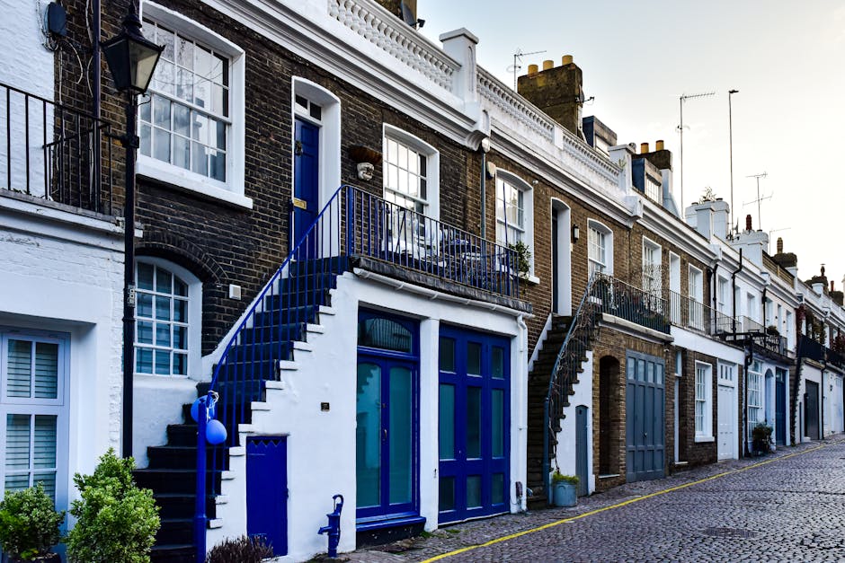 A row of traditional brick terraced houses on Brompton Road with white window frames, black and white painted external stairs, and blue doors and railings. The cobblestone street in front appears clean and well-maintained with some small green shrubs near the house entrance. The scene is illuminated by natural daylight, highlighting details of the external surfaces and the neat appearance of the facades. Carpet Cleaners SW7 is mentioned as the service provider for domestic cleaning and surface sanitisation in the SW7 area, emphasizing thorough deep cleaning and maintenance of residential exteriors.