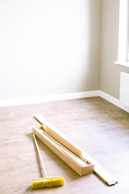 A bright, minimally furnished room with light-colored wooden flooring and beige walls, illuminated by natural light from a large window on the right. On the floor, there is a wooden window frame with a sliding glass pane, a long-handled yellow cleaning brush placed nearby, and a narrow, flat wooden strip. The room appears clean and well-maintained, with no visible dust or clutter, showcasing a surface cleaning or preparatory stage for deep cleaning or maintenance. The image reflects the scheduling or ongoing process of window frame and floor surface cleaning, typical of domestic or commercial sanitisation and cleaning tasks. The setting is associated with professional cleaning services provided by Carpet Cleaners SW7, as indicated by the page title about end of tenancy cleaning tips and costs on the website.
