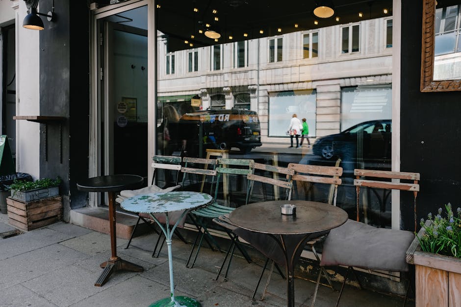The image depicts the exterior of a commercial establishment on Kensington High Street, with large glass windows reflecting the street scene, including a black vehicle and pedestrians. The outdoor seating area features a variety of tables and chairs with different materials: a small round table with a floral-patterned tabletop, a dark wooden table with an ashtray on top, and a few wooden and metal chairs, some with light-colored cushions. Potted plants and greenery are positioned on the pavement near the seating area, contributing to a welcoming atmosphere. The lighting suggests daytime, with natural light illuminating the scene, highlighting the cleanliness and tidiness of the outdoor space. Elements such as the glass, wood, and metal surfaces appear well-maintained and free of dust or debris, aligning with professional surface cleaning practices promoted by Carpet Cleaners SW7, especially relevant to upholstery and surface sanitisation for shops and commercial spaces as detailed in the Kensington High Street upholstery cleaning guide for shops, SW7.