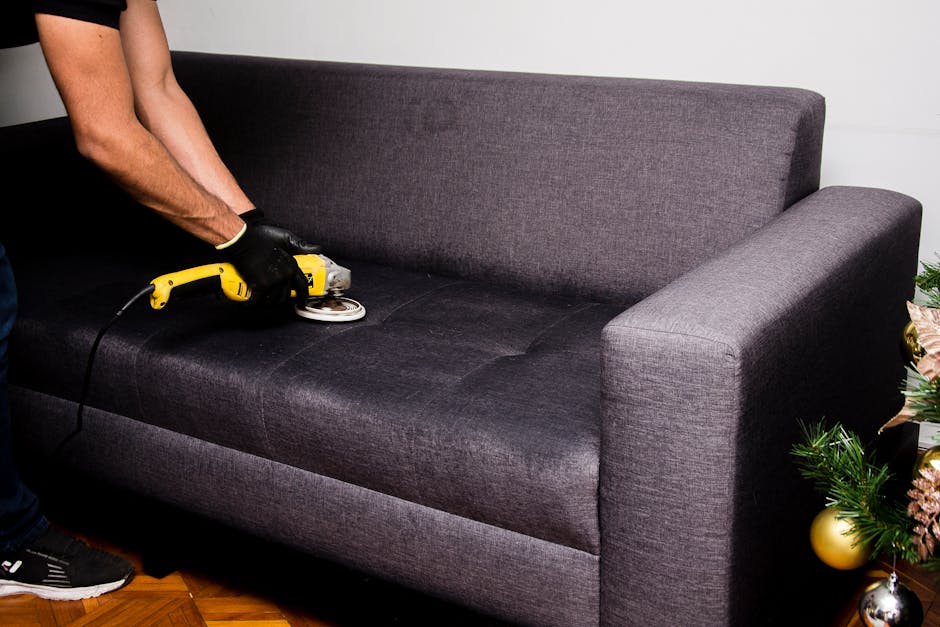 A person wearing black gloves is using a handheld electric upholstery steamer or steam cleaning tool to deep clean a dark grey fabric sofa in a living room setting. The sofa has a smooth, tidy appearance with visible upholstery surface, and the person is focusing on an area of the seat cushions. The background includes a white wall and a small festive decoration with green foliage and gold ornaments placed to the right of the sofa. The room is well-lit, showcasing a clean and maintained environment. This surface cleaning aligns with the services provided by Carpet Cleaners SW7, highlighting professional upholstery cleaning and sanitisation for residential spaces on Kensington High Street.