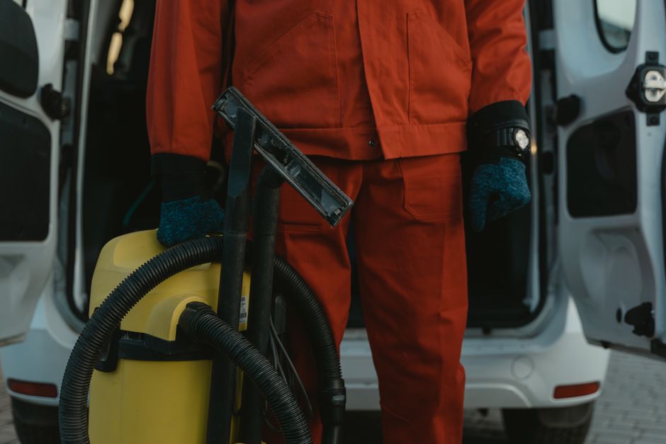 A professional cleaner wearing a blue uniform and a yellow cleaning bucket with tools stands outside a modern building with large glass windows, performing surface cleaning and window sanitisation. Th