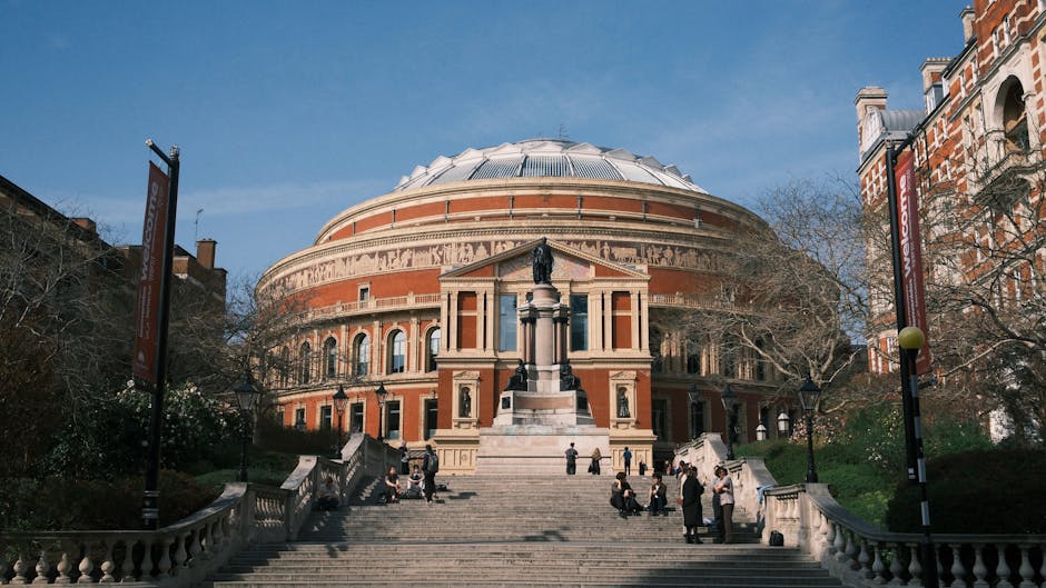 The exterior view of the Royal Albert Hall showing a large, circular, domed brick building with tall arched windows and decorative stone accents, set against a clear blue sky. In the foreground, a wide stone staircase leads up to the entrance, where visitors are sitting and standing on the steps. A statue on a pedestal is positioned at the top of the staircase, surrounded by green grassy areas with trees and shrubs on either side. The scene is well-lit with natural daylight, emphasizing the building's historic architecture and clean surroundings, consistent with professional cleaning and maintenance standards as highlighted by Carpet Cleaners SW7.