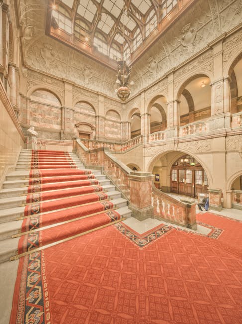 The exterior view of the Royal Albert Hall showing a large, circular, domed brick building with tall arched windows and decorative stone accents, set against a clear blue sky. In the foreground, a wide stone staircase leads up to the entrance, where visitors are sitting and standing on the steps. A statue on a pedestal is positioned at the top of the staircase, surrounded by green grassy areas with trees and shrubs on either side. The scene is well-lit with natural daylight, emphasizing the building's historic architecture and clean surroundings, consistent with professional cleaning and maintenance standards as highlighted by Carpet Cleaners SW7.