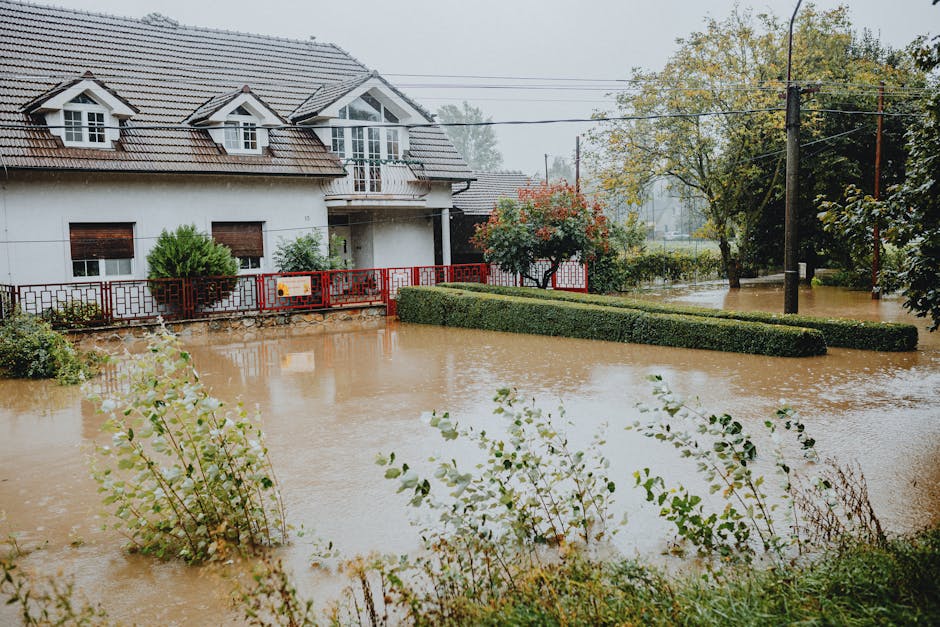 A residential property during a flood, with the front yard submerged under muddy water. The house features a white exterior with a gabled roof covered in grey tiles, and several windows with white frames. A red metal fence surrounds the property, partially obscured by the high water level. The front garden has neatly trimmed bushes and small trees, some partially submerged. Overcast skies and dense trees in the background suggest rainy weather contributing to the flood. The scene captures the extent of flood damage and highlights the importance of professional surface cleaning and flood damage restoration services offered by Carpet Cleaners SW7, who provide same-day emergency responses in SW7 to restore hygiene and safety to affected properties.