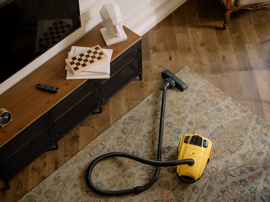 A yellow handheld vacuum cleaner with a flexible hose and wand attachment resting on a patterned area rug in a living room. The vacuum is positioned near a wooden media unit with black metal mesh doors, which holds a stack of papers and a chessboard, with a white sculptural bust and a remote control on top. The room has wooden flooring and a white skirting board along the wall, with soft natural light illuminating the space and highlighting the cleanliness of the surfaces. This image reflects surface cleaning and domestic deep cleaning processes, showcasing a tidy and well-maintained living area as part of routine hygiene and surface maintenance, with Carpet Cleaners SW7 providing professional cleaning solutions.