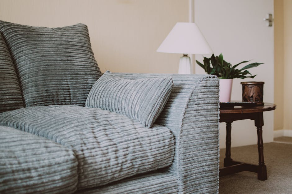 A close-up view of a textured grey fabric sofa with corduroy upholstery, situated in a living room with neutral-colored walls. The sofa is clean and well-maintained, featuring a matching cushion. To t