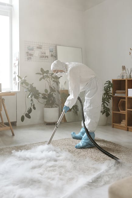 Close-up of a person using a handheld carpet cleaning machine on a textured beige rug in a bright, modern room with white walls. The individual is barefoot, wearing light-colored pants and a yellow to