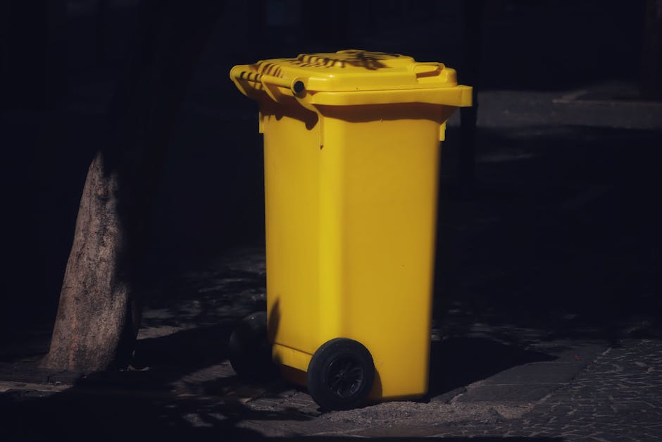 A yellow wheeled waste bin with a closed lid is positioned outdoors on a paved surface, partially shaded by shadows. The bin is situated next to a stone curb and a rock, with minimal surrounding elements visible in the darkened environment. The surface beneath the bin appears clean, and the image emphasizes the cleanliness and maintenance of waste disposal containers, relating to hygienic practices in residential or commercial cleaning, consistent with surface cleaning and waste management topics discussed on the page about Kensington and Chelsea council disposal rules. The scene is lit with natural or ambient lighting, highlighting the bright yellow color of the bin against the darker background, and is associated with the services provided by Carpet Cleaners SW7.