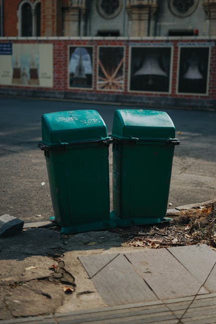 A yellow wheeled waste bin with a closed lid is positioned outdoors on a paved surface, partially shaded by shadows. The bin is situated next to a stone curb and a rock, with minimal surrounding elements visible in the darkened environment. The surface beneath the bin appears clean, and the image emphasizes the cleanliness and maintenance of waste disposal containers, relating to hygienic practices in residential or commercial cleaning, consistent with surface cleaning and waste management topics discussed on the page about Kensington and Chelsea council disposal rules. The scene is lit with natural or ambient lighting, highlighting the bright yellow color of the bin against the darker background, and is associated with the services provided by Carpet Cleaners SW7.